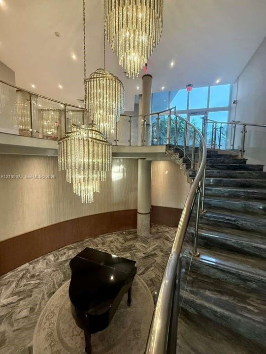 Grand staircase featuring stone-finish steps, a curved glass railing with metal accents, tiered crystal chandeliers, and patterned stone flooring
