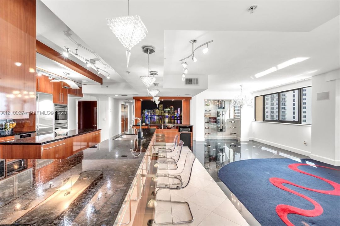 Contemporary kitchen featuring high-gloss wood-finish cabinetry, expansive stone countertops, integrated stainless steel appliances, a tiered island with built-in seating, and intricate custom overhead lighting