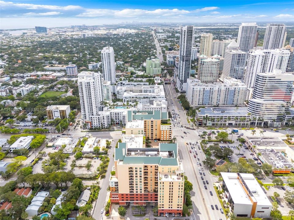 Aerial perspective of a multi-story urban complex featuring a prominent peaked roofline, stucco-finish facade, ground-level parking access, and extensive perimeter greenery