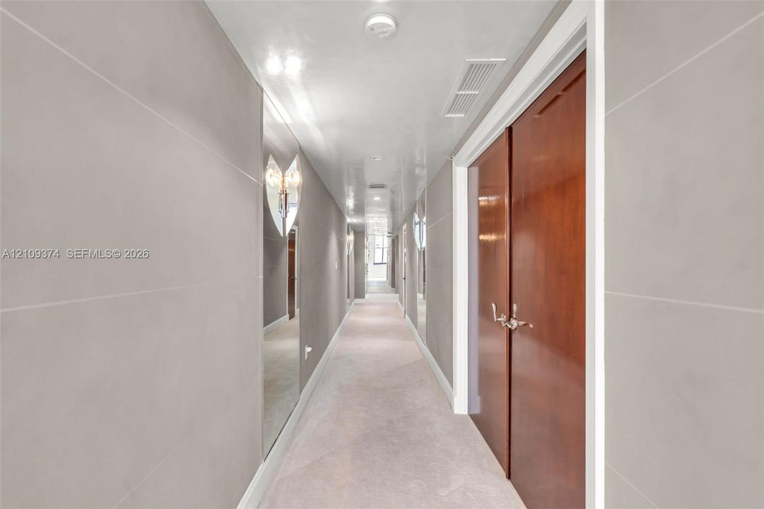 Hallway featuring large-format light gray wall panels, full-height wall mirrors, double-door wood cabinetry, and recessed lighting