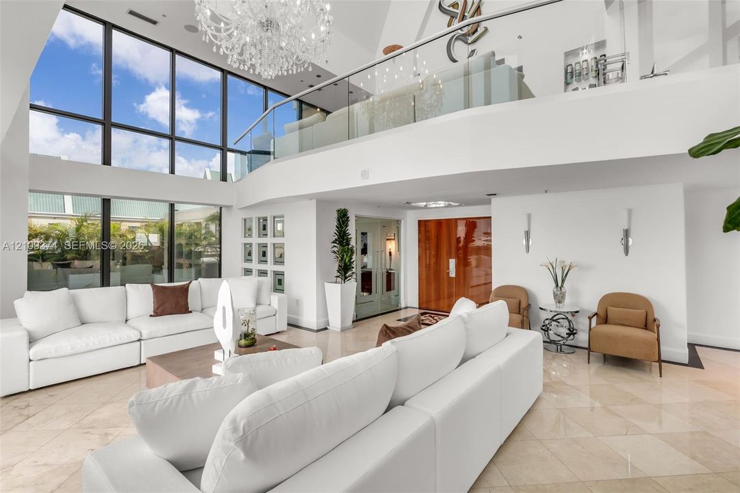 Double-height living area featuring floor-to-ceiling windows, polished stone flooring, a glass mezzanine railing, and a grand crystal chandelier