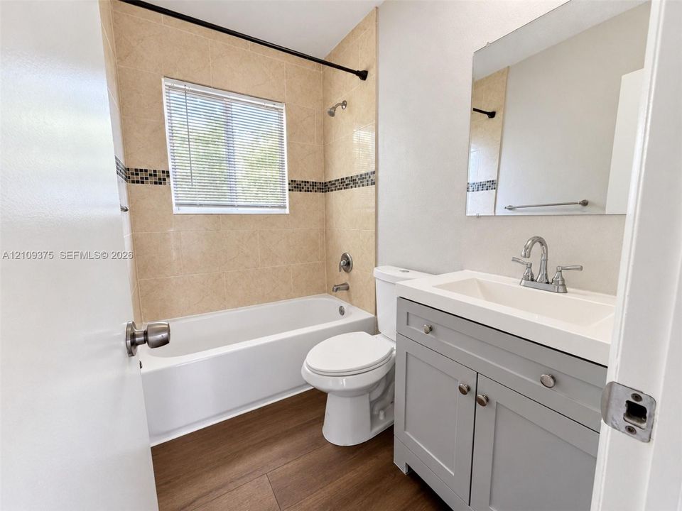 Bathroom featuring wood-finish flooring, a grey vanity with an integrated basin, a soaking tub with tiled surround and decorative accent border, and a wall-mounted mirror