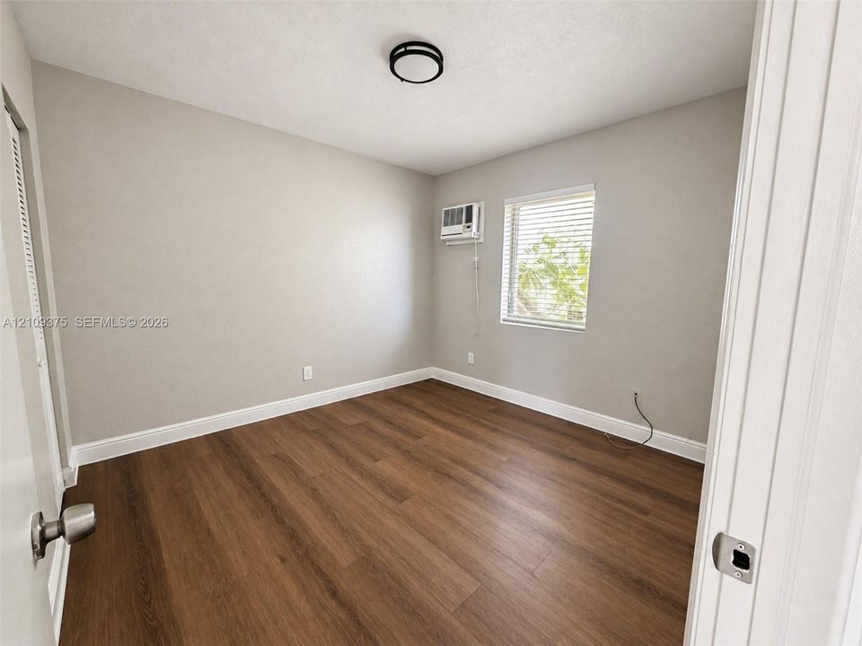 Room featuring wood-finish flooring, a neutral wall color, a single-window configuration with horizontal blinds, a wall-mounted air conditioning unit, and a flush-mount circular ceiling light