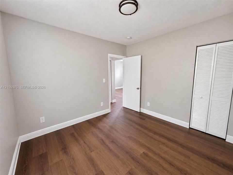 Neutral-toned interior room featuring wood-finish flooring, a white six-panel door, and a built-in closet with white louvered doors, completed by a flush-mount circular ceiling light