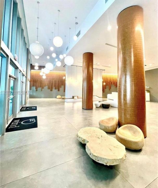 Grand lobby entrance featuring high ceilings, floor-to-ceiling windows, polished stone flooring, custom pendant globe lighting, and textured metallic-finish structural columns