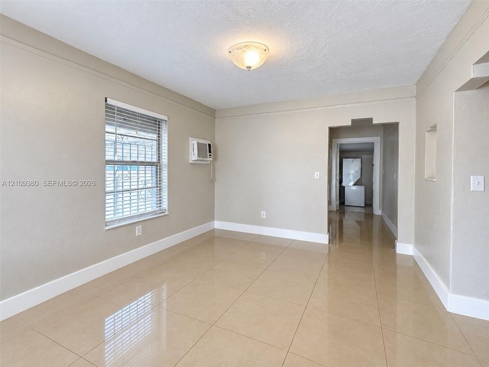 Neutral-toned interior space featuring large-format tile flooring, a window with horizontal blinds, a wall-mounted climate control unit, and a flush-mount ceiling light