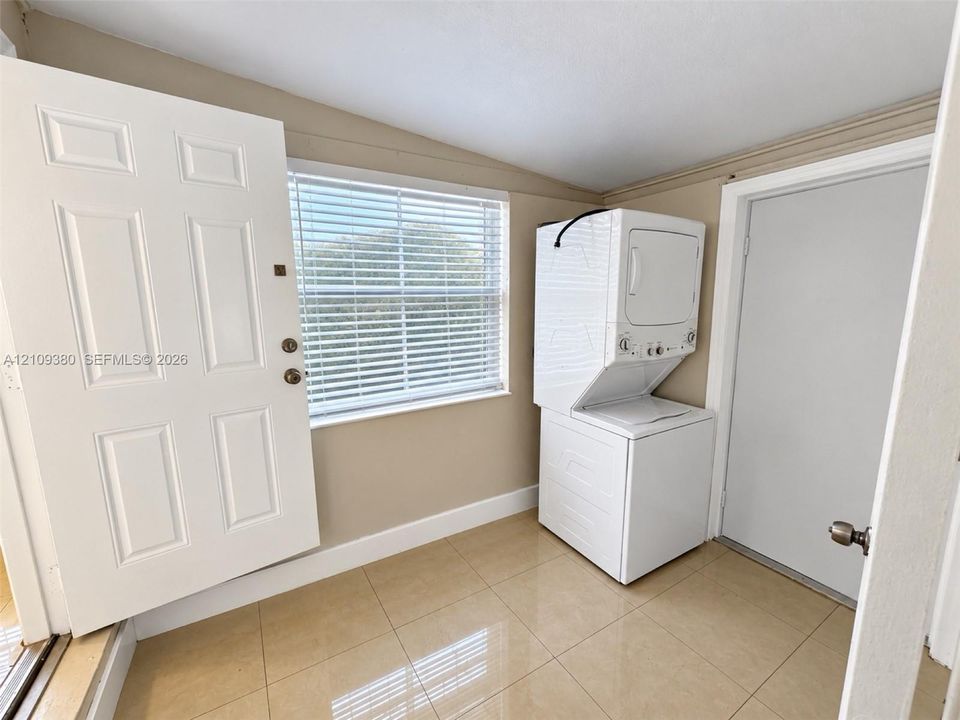 Laundry area featuring neutral tile flooring, a window with horizontal blinds, and a stacked washer and dryer unit