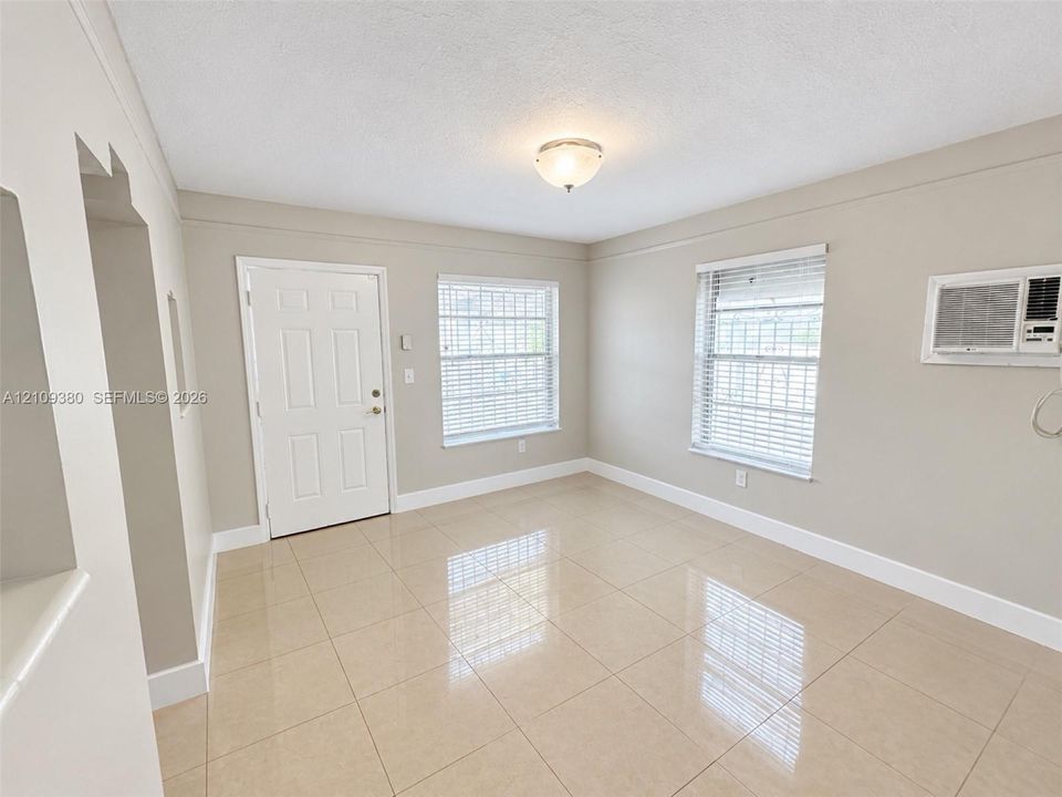 Neutral-toned interior featuring large-format ceramic tile flooring, two multi-pane windows, a flush-mount ceiling light fixture, and a wall-mounted air conditioning unit