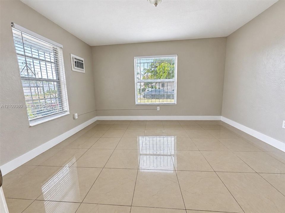 Neutral-toned interior space featuring polished tile flooring, dual windows with integrated security bars, and a wall-mounted cooling unit