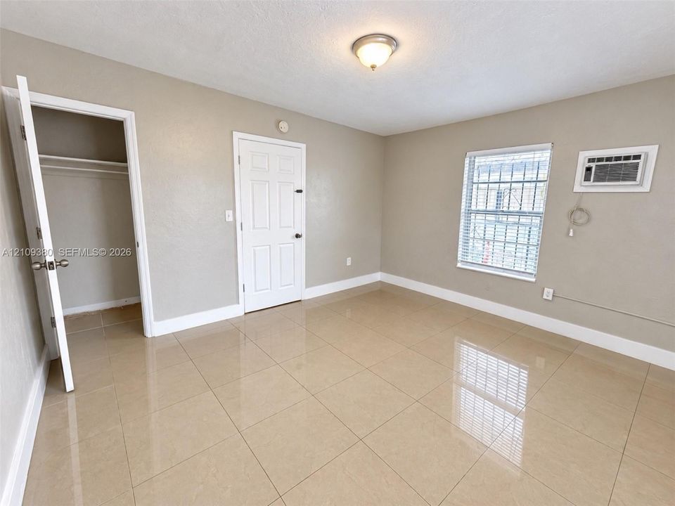 Neutral-toned interior featuring large-format ceramic tile flooring, a recessed closet with built-in shelving, a six-panel door, a window with metal security bars, and a wall-mounted air conditioning unit