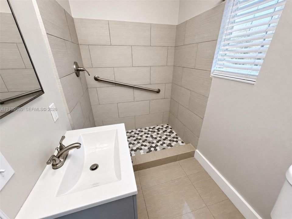 Wall-mounted sink with a double-handle faucet, neutral tile shower surround, hexagonal mosaic floor tiling, metal support bar, and horizontal blinds