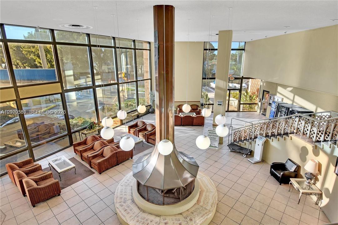 Floor-to-ceiling multi-pane windows, central cylindrical fireplace with metallic finish, globe pendant lighting, ceramic tile flooring, and decorative metal stair railing