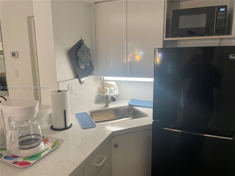 Kitchenette featuring glossy white cabinetry, solid-surface countertops, an undermount stainless steel sink with a goose-neck faucet, and a black full-size refrigerator
