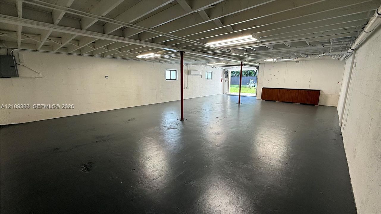 Open-concept utility space featuring painted masonry walls, sealed concrete flooring, exposed ceiling joists, linear fluorescent lighting, and built-in cabinetry