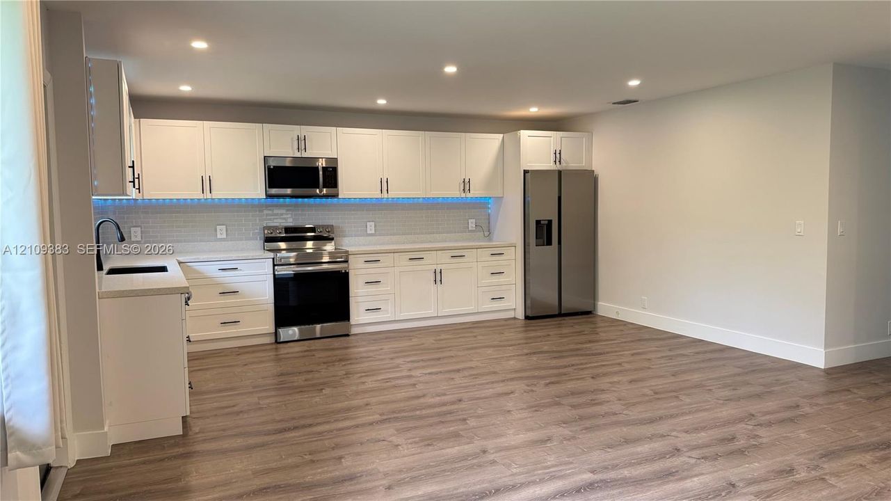 Kitchen featuring white shaker cabinetry, stainless steel appliances, tile backsplash with integrated lighting, quartz countertops, and wood-finish flooring