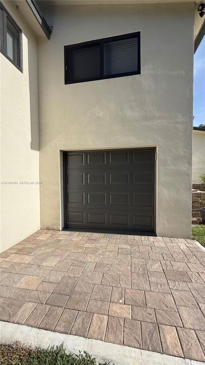 Stucco exterior featuring a single-car garage with a paneled door, a rectangular window with a dark frame, and a patterned paver driveway