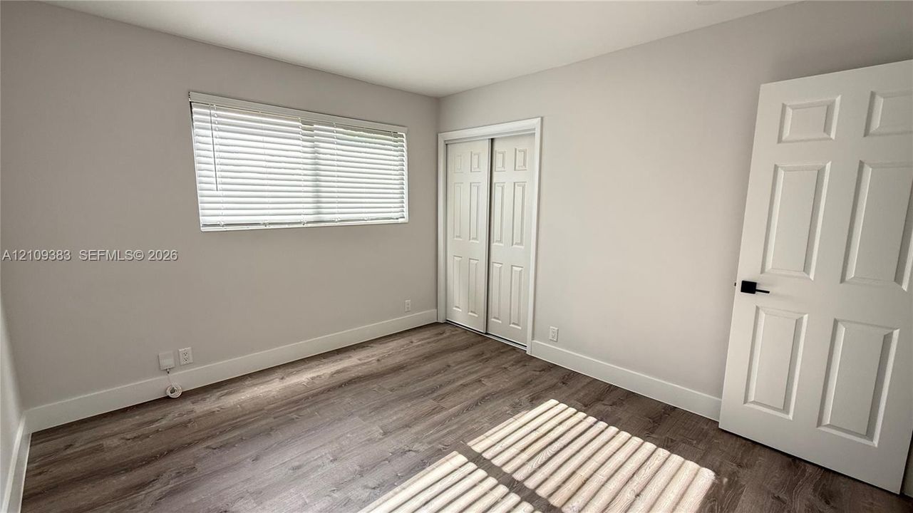 Room featuring wood-finish flooring, neutral wall paint, a sliding-door closet, a six-panel interior door with matte black hardware, and an installed window blind