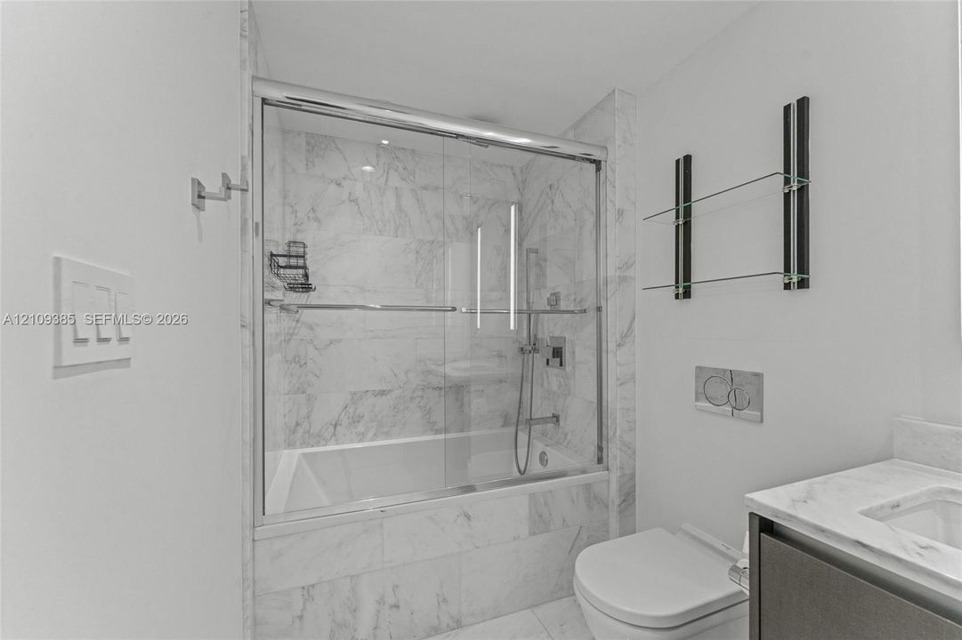 Wall-mounted dark wood-finish vanity with stone countertop, integrated sink, LED-lit mirror, built-in glass shelving, and wall-mounted dual-flush actuator