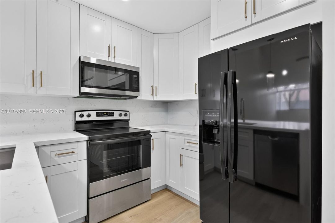 Kitchen featuring white shaker-style cabinetry with gold-tone hardware, white stone-composite countertops, wood-finish flooring, and black-finish stainless steel appliances