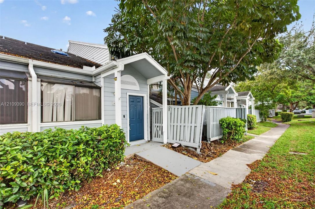 Light blue siding, covered entryway with gabled roof, blue paneled front door, and private fenced patio