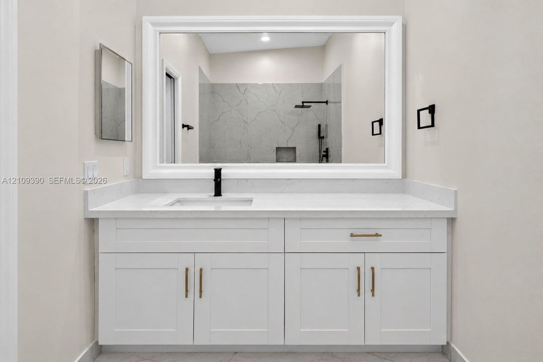 White shaker-style cabinetry with brass hardware, solid surface countertop, rectangular undermount sink, matte black faucet, and large framed wall mirror