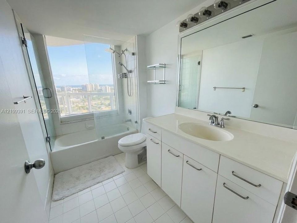 Bathroom featuring a white vanity with integrated countertop sink, full-width wall mirror, shower-tub combination with glass enclosure, and large-format window