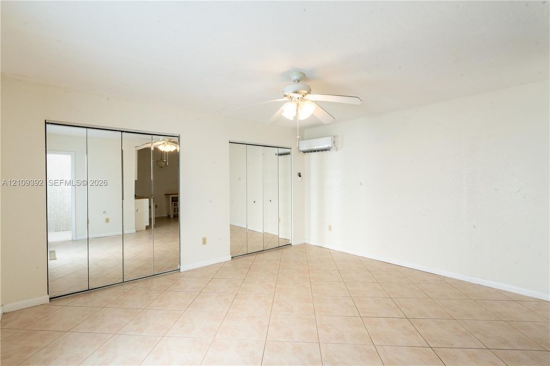Neutral-toned tile flooring, dual mirrored sliding-door closets, ceiling fan with integrated lighting, and wall-mounted climate control unit