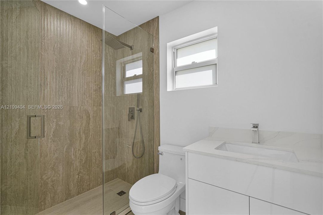 Bathroom featuring a walk-in shower with stone-tile surround, glass enclosure, and rainfall showerhead, complemented by a white floating vanity with a stone countertop and integrated sink
