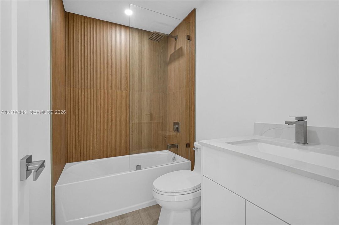 Bathroom featuring a tub with a floor-to-ceiling slatted wood-finish surround, a glass panel shower guard, a white vanity with an integrated basin, and brushed nickel hardware