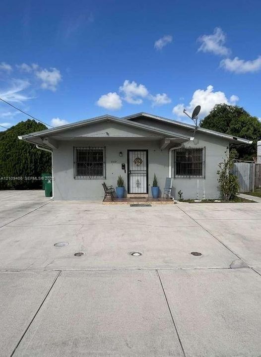 Single-story exterior featuring a concrete facade, gabled roofline, metal security window grates, and an expansive concrete parking area
