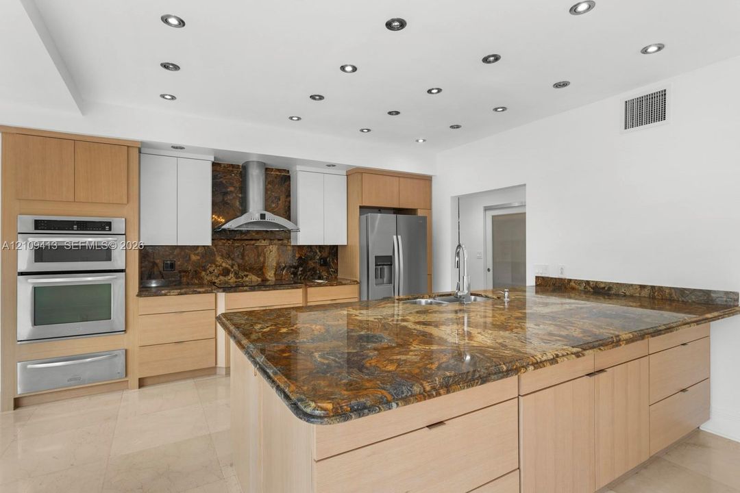 Kitchen featuring custom wood-finish cabinetry, stone slab countertops, a central island with an integrated sink, built-in double wall ovens, and recessed ceiling lighting