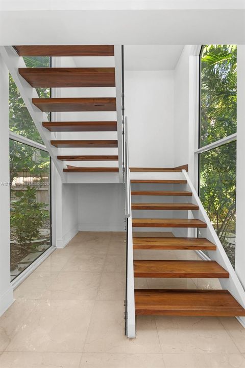 Open-riser staircase featuring wood-finish treads, white structural stringers, a sleek metal handrail, floor-to-ceiling glass windows, and light-toned tile flooring