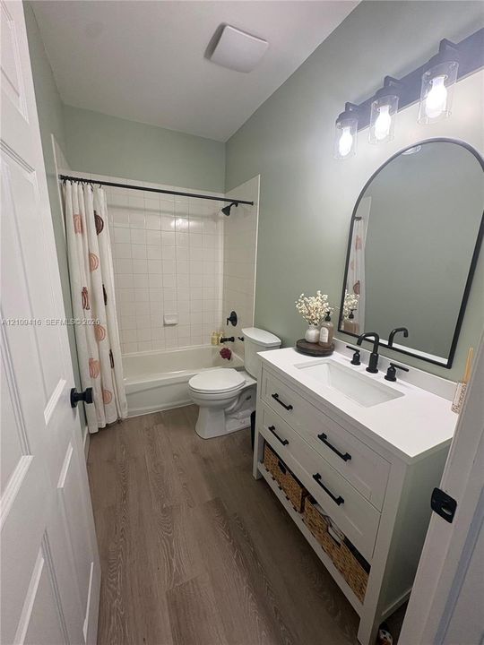 Bathroom featuring a white vanity with a rectangular sink, arch-top mirror, matte black wall-mounted light fixture, wood-finish flooring, and a tub-shower combination with tile surround