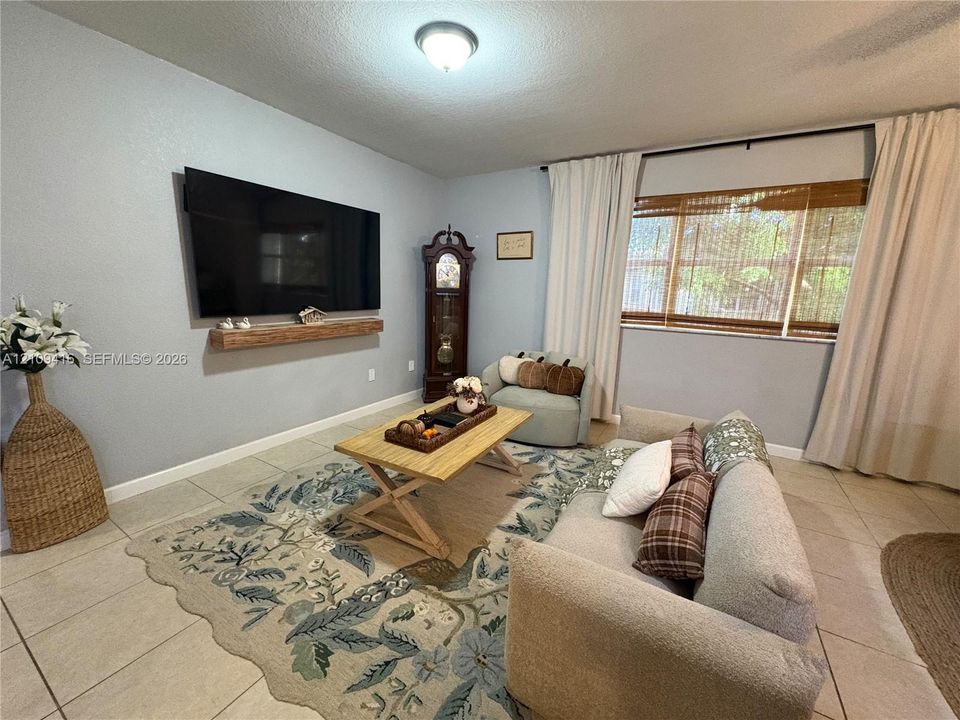 Neutral-toned living space featuring large-format tile flooring, a wall-mounted floating wood mantel, and windows finished with integrated blinds and full-length drapery