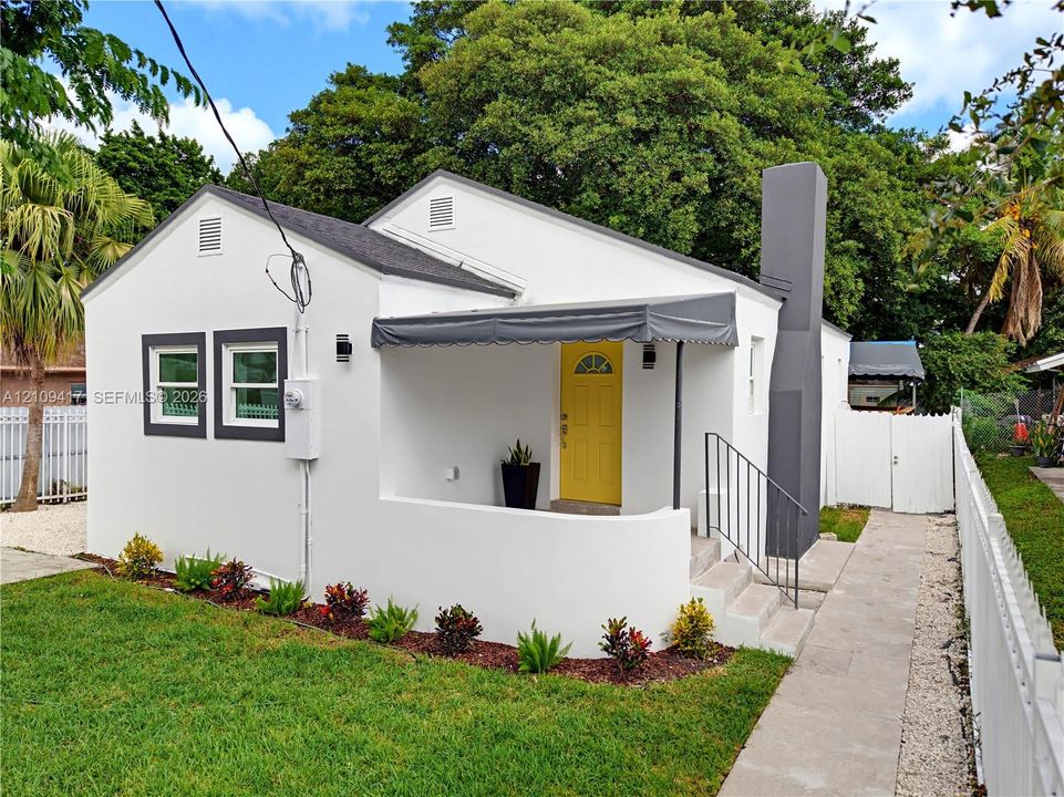 Exterior featuring a stucco facade, covered front entry with a metal awning, prominent exterior chimney, and a white perimeter fence