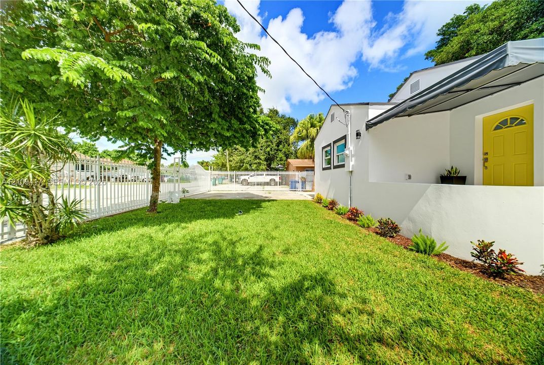 Side yard featuring a grass lawn, metal perimeter fencing, mature trees, concrete driveway, and an exterior awning over the entrance