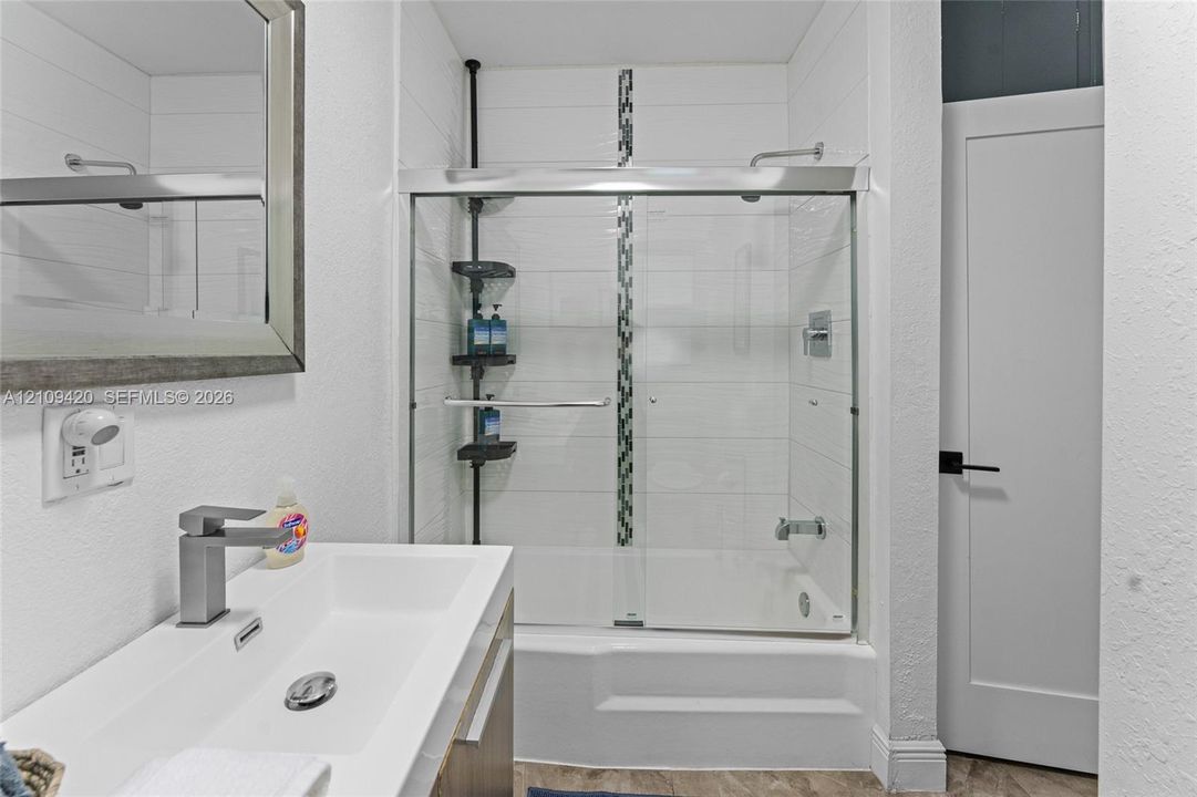 Modern vanity with a rectangular sink, wall-mounted mirror, glass-enclosed shower and tub combination featuring horizontal tile surrounds and a decorative mosaic accent strip