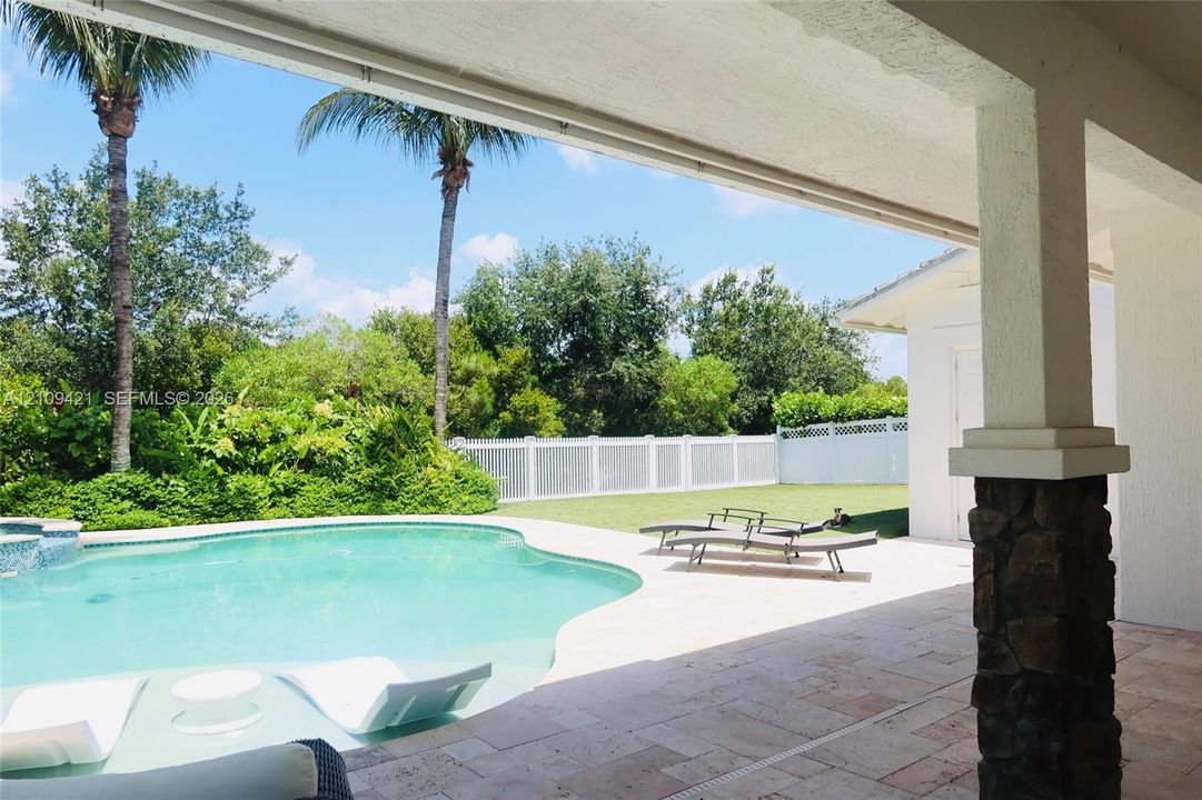 Covered patio with natural stone flooring and stone-accented pillar, swimming pool with built-in tanning ledge, and white vinyl privacy fencing