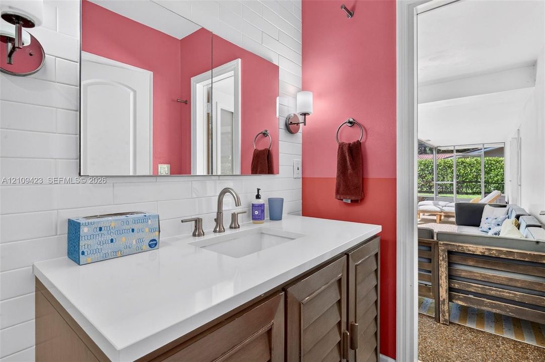 Vanity featuring a solid-surface countertop with an integrated basin, shutter-style wooden cabinetry, horizontal subway tile backsplash, wall-mounted lighting, and a wide-format mirror