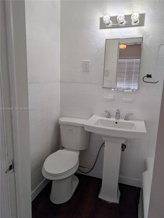 White pedestal sink featuring a chrome faucet, wall-mounted rectangular mirror, white square tile wainscoting, and wood-finish flooring