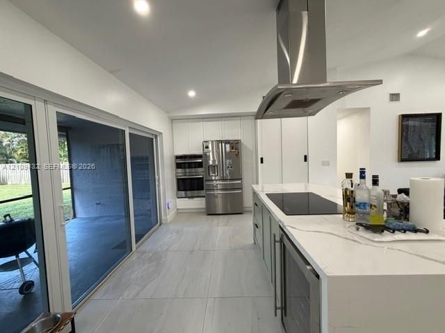 Kitchen featuring a large waterfall-edge island with an integrated cooktop and under-counter refrigeration, stainless steel wall-mounted range hood, custom cabinetry, stainless steel double wall ovens, and expansive sliding glass doors