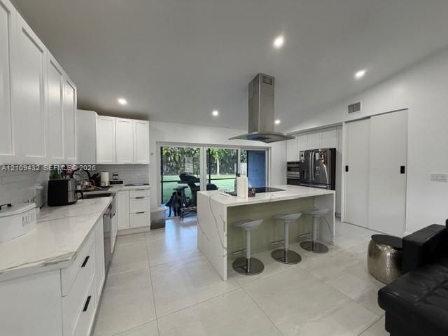 Kitchen featuring white cabinetry, waterfall-edge island with stone-finish countertop, stainless steel vent hood, recessed lighting, and large-format tile flooring