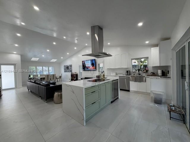 Open-concept interior featuring vaulted ceilings, oversized kitchen island with stone-finish countertops, stainless steel chimney-style range hood, farmhouse sink, and large-format tile flooring