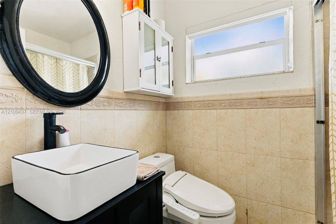 Rectangular vessel sink with matte black faucet, dark vanity cabinet, circular mirror, wall-mounted storage cabinetry, and tiled wall wainscoting