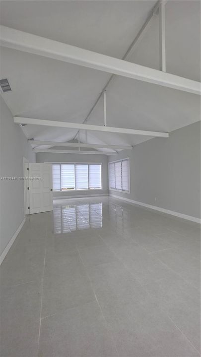 Vaulted ceiling with exposed white beams, large-format tile flooring, and multiple windows with horizontal blinds