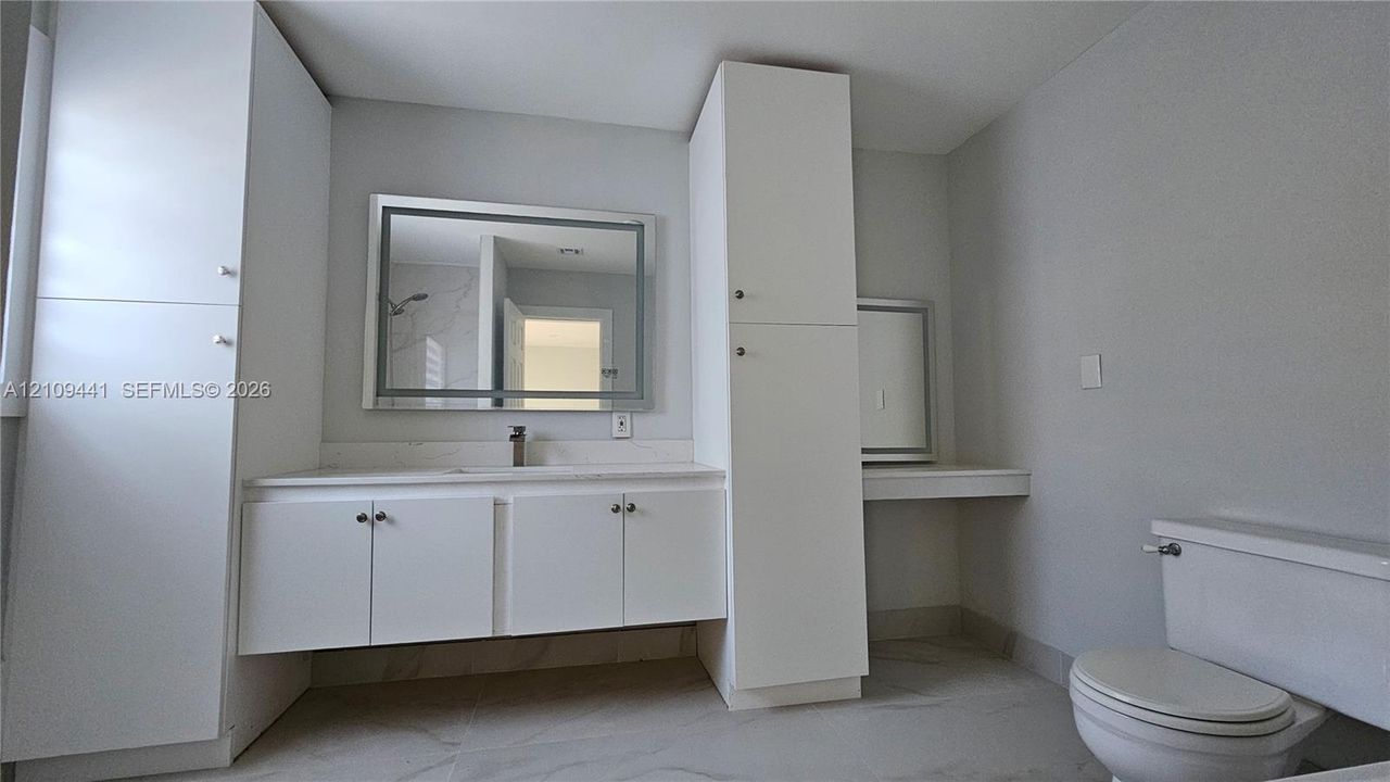 Bathroom featuring white cabinetry with integrated floor-to-ceiling storage, stone-surface countertop, rectangular wall mirror with integrated lighting, and neutral large-format tile flooring