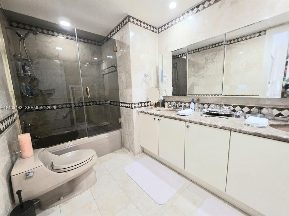 Bathroom featuring stone-tiled walls with decorative mosaic trim, dual-sink vanity with solid-surface countertop, frameless glass shower enclosure, and recessed lighting