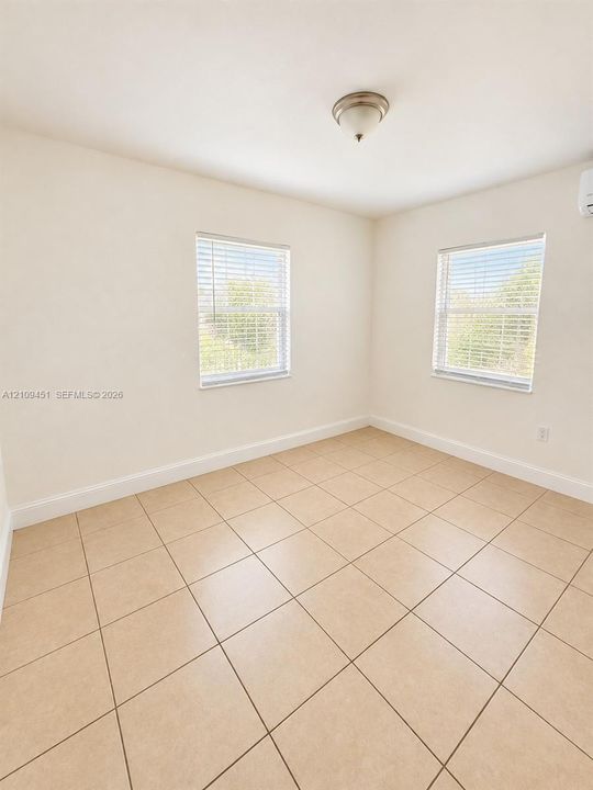 Interior space featuring large-format ceramic tile flooring, neutral wall tones, two windows with horizontal blinds, and a wall-mounted climate control unit