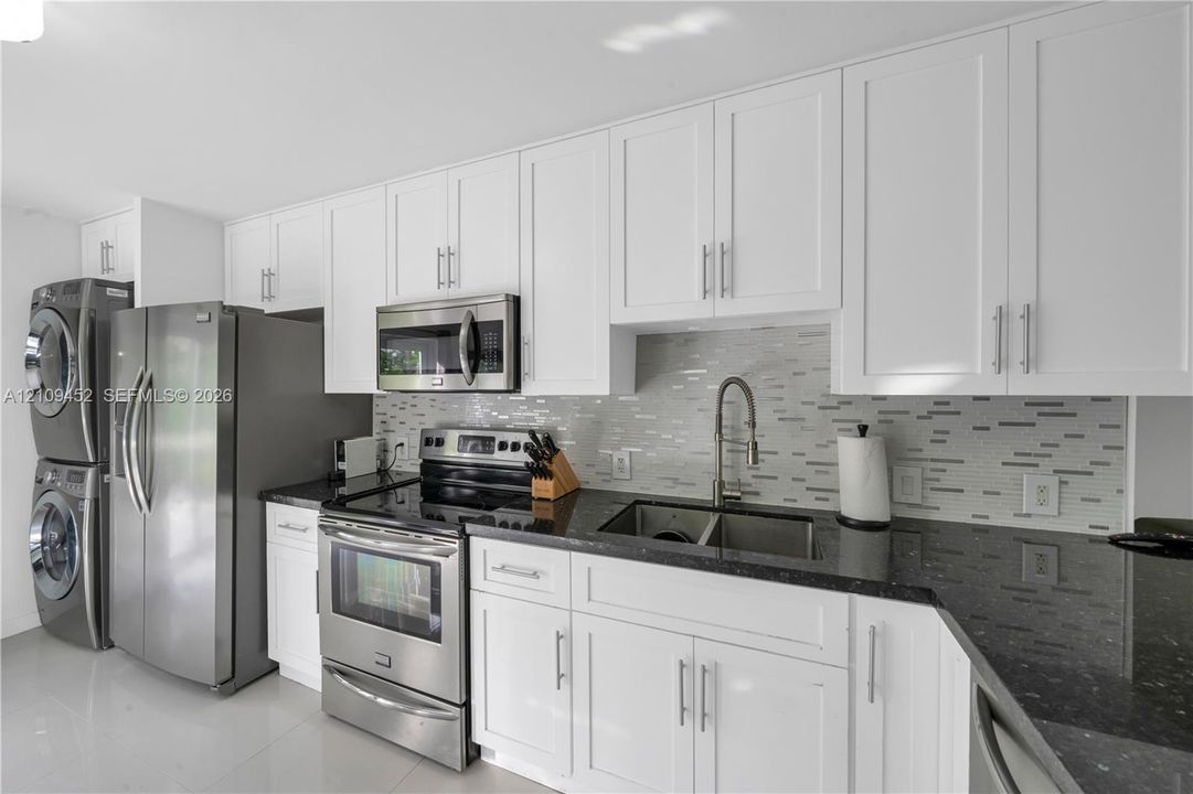 Kitchen featuring white shaker-style cabinetry, dark stone countertops, a mosaic tile backsplash, stainless steel appliances, and an integrated laundry area