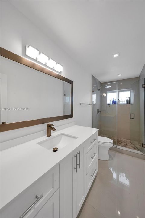 White vanity with a solid-surface countertop and integrated sink, bronze-tone faucet and mirror frame, multi-light vanity fixture, and a glass-enclosed shower with tiled flooring